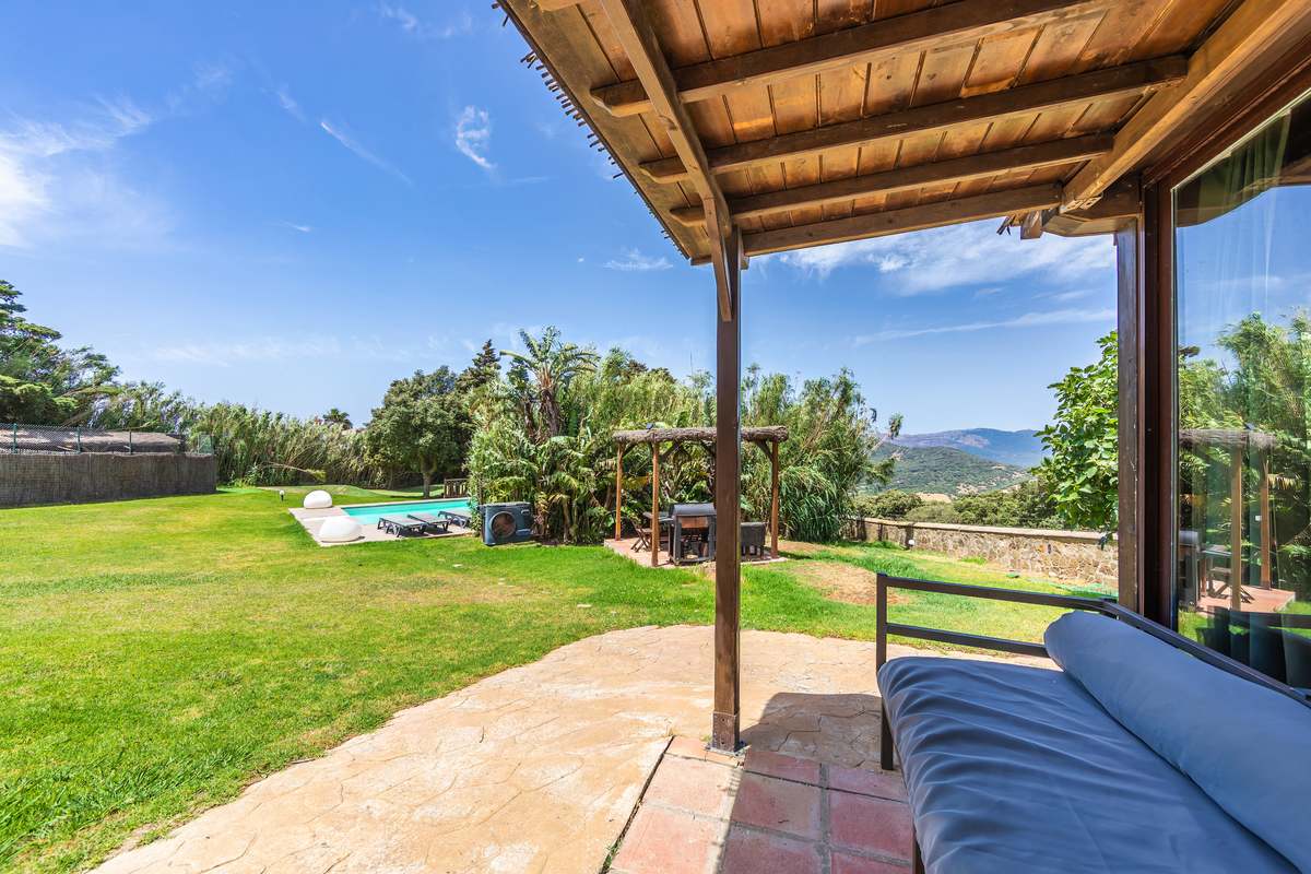 Porche cubierto con sofá azul y vistas al jardín, piscina y montañas en un alojamiento de Tarifa