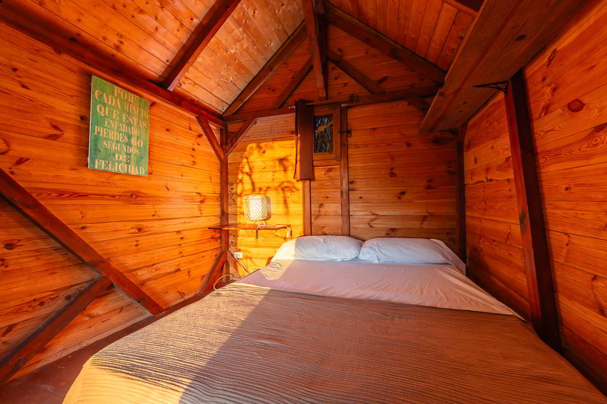 Cama doble dentro de la cabaña del Chalet La Ahumada, con paredes de madera, lámpara de mimbre encendida y luz cálida de atardecer.