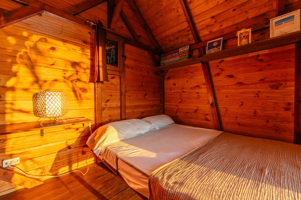 Cama doble en el rincón de la cabaña del Chalet La Ahumada, con lámpara encendida, detalles decorativos y cálida luz del atardecer.