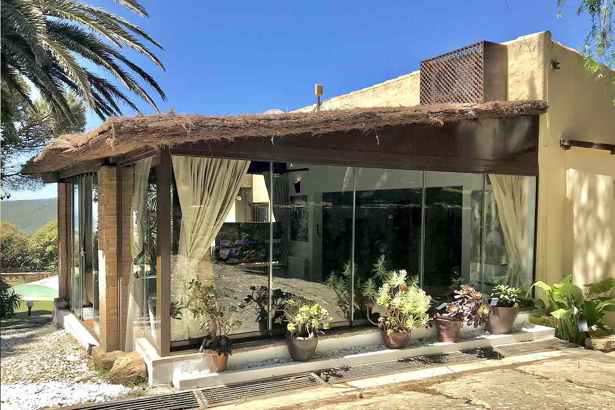 Fachada acristalada del chalet La Ahumada, rodeada de plantas y palmeras, con acceso directo al jardín.