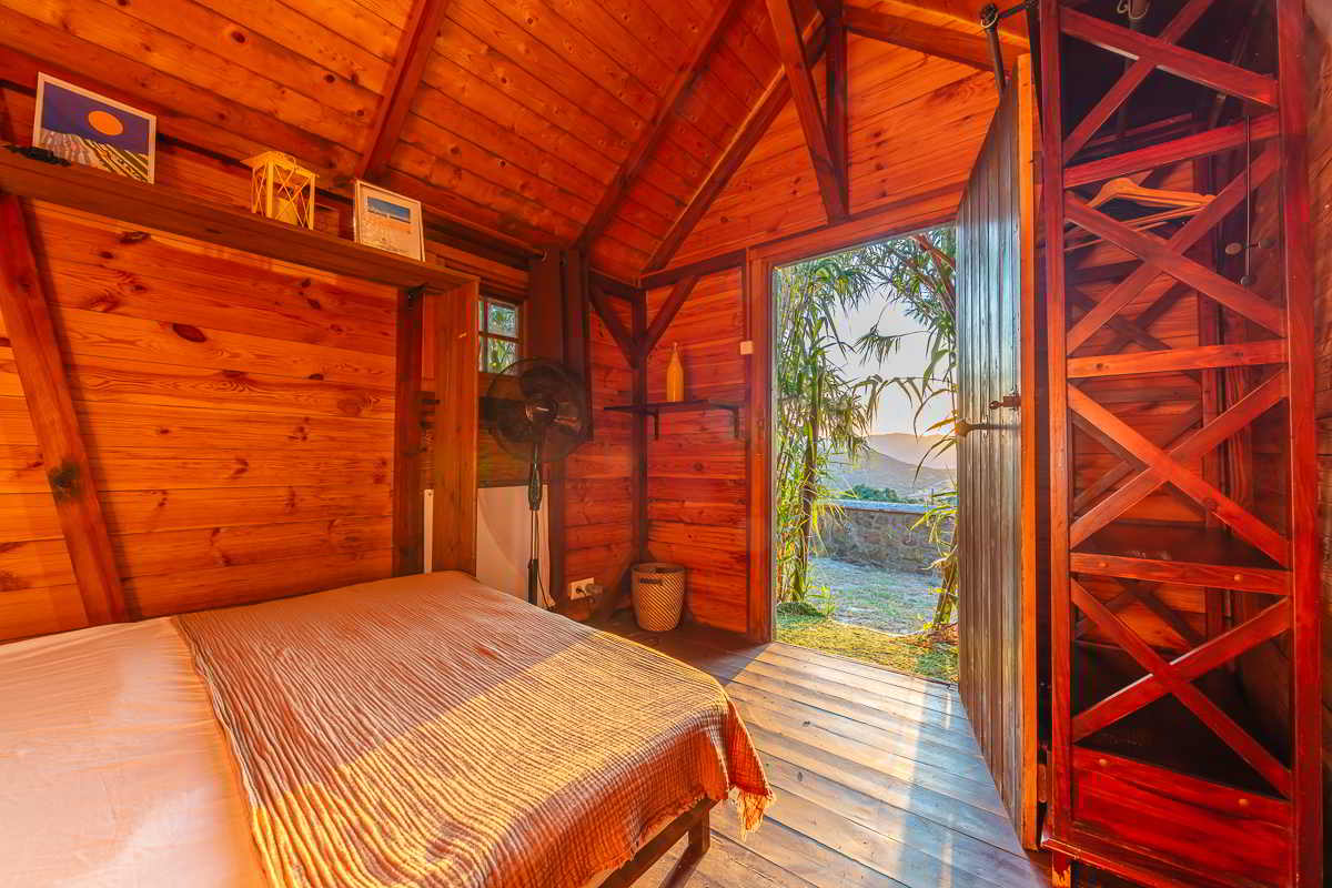 Interior de la cabaña del Chalet La Ahumada con cama doble, armario abierto de madera, ventilador y puerta abierta hacia el campo.