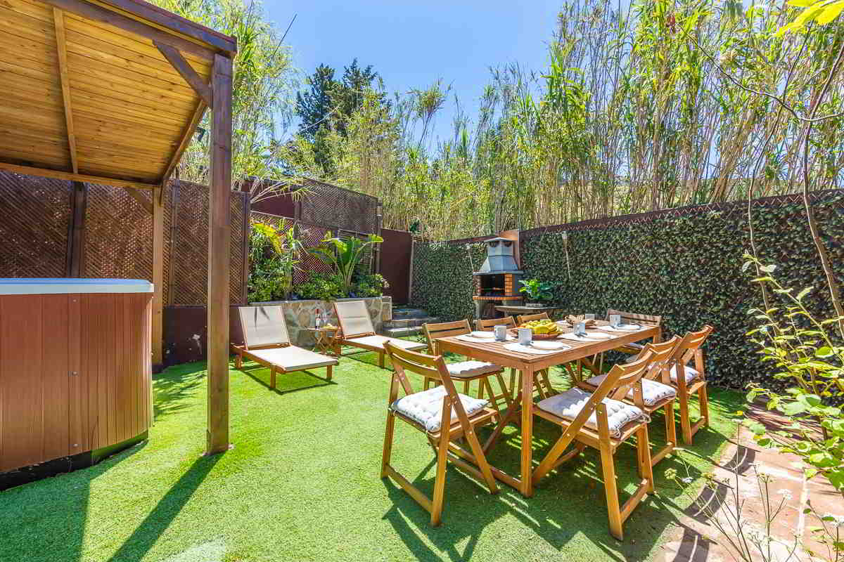 Jardín de Villa Verde en Tarifa con zona de comedor exterior de madera, barbacoa de obra, tumbonas y parte de la piscina cubierta al fondo, rodeado de vegetación alta y muros naturales.