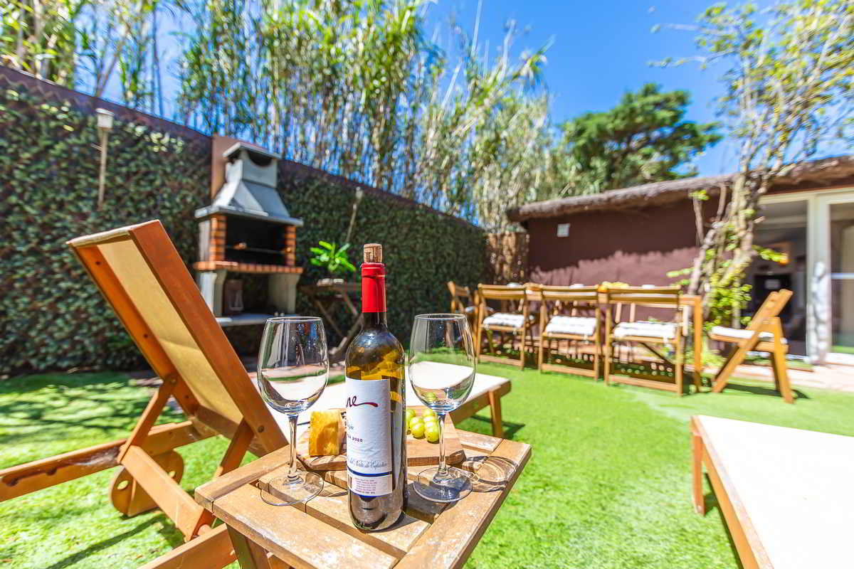 Jardín de Villa Verde en Tarifa con zona de comedor exterior de madera, barbacoa de obra, tumbonas y parte de la piscina cubierta al fondo, rodeado de vegetación alta y muros naturales.