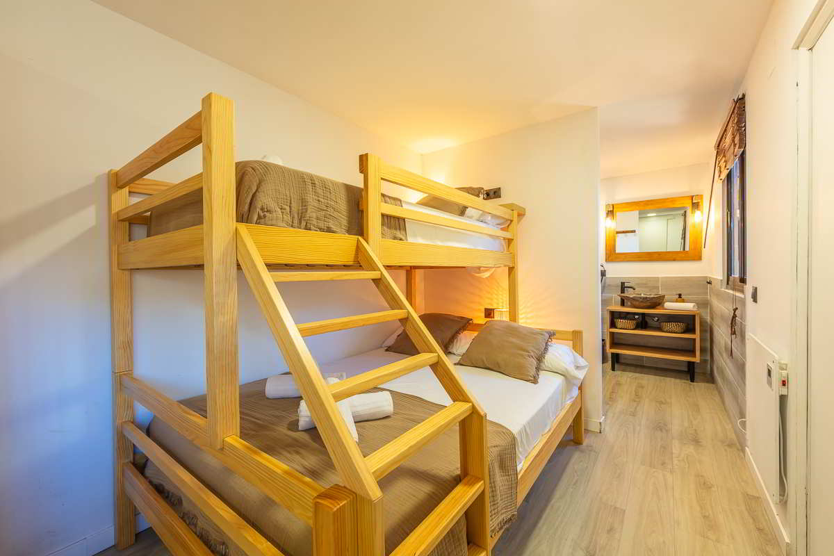 Cama litera de madera con colchón doble en la parte inferior y colchón individual en la parte superior, con ropa de cama en tonos marrones y almohadas decorativas, vista parcial del baño con lavamanos de piedra y espejo de madera al fondo.
