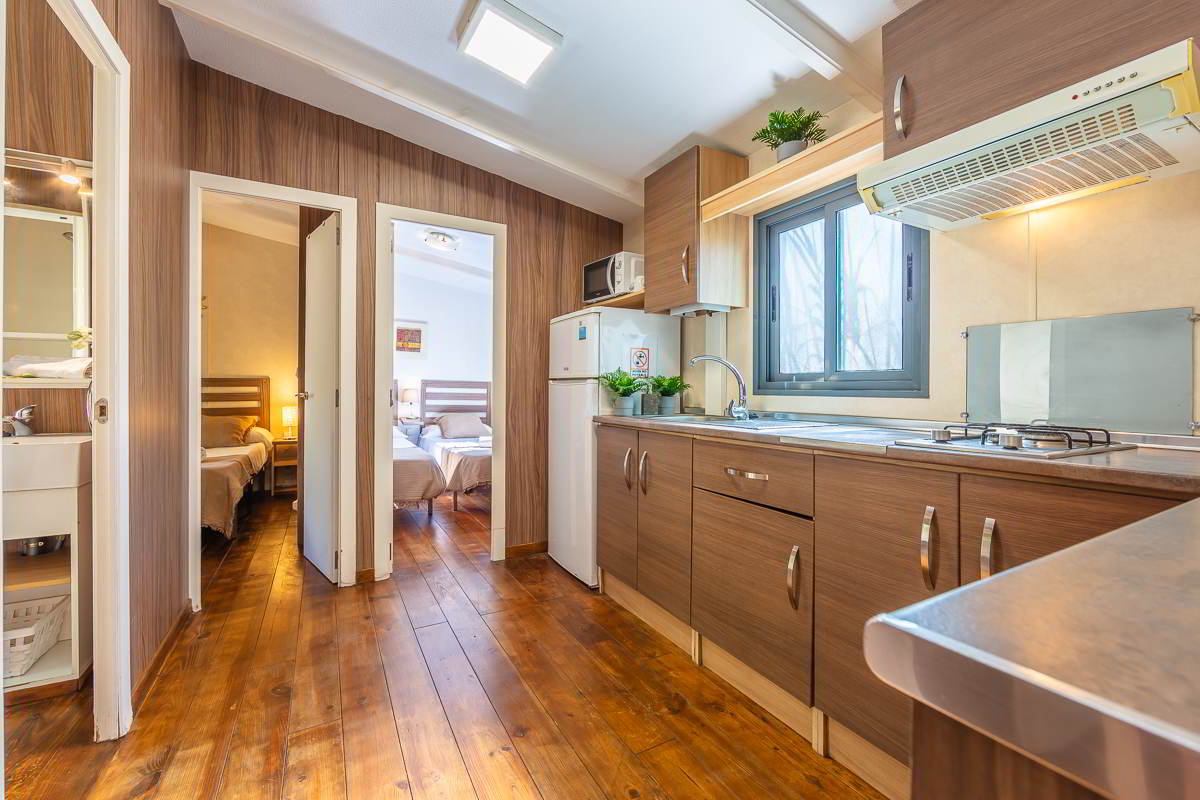 Cocina de la casa Nepal en Villas La Ahumada, con muebles de madera, encimera de acero, ventana con luz natural, frigorífico, microondas y acceso a dos dormitorios con camas individuales y baño lateral