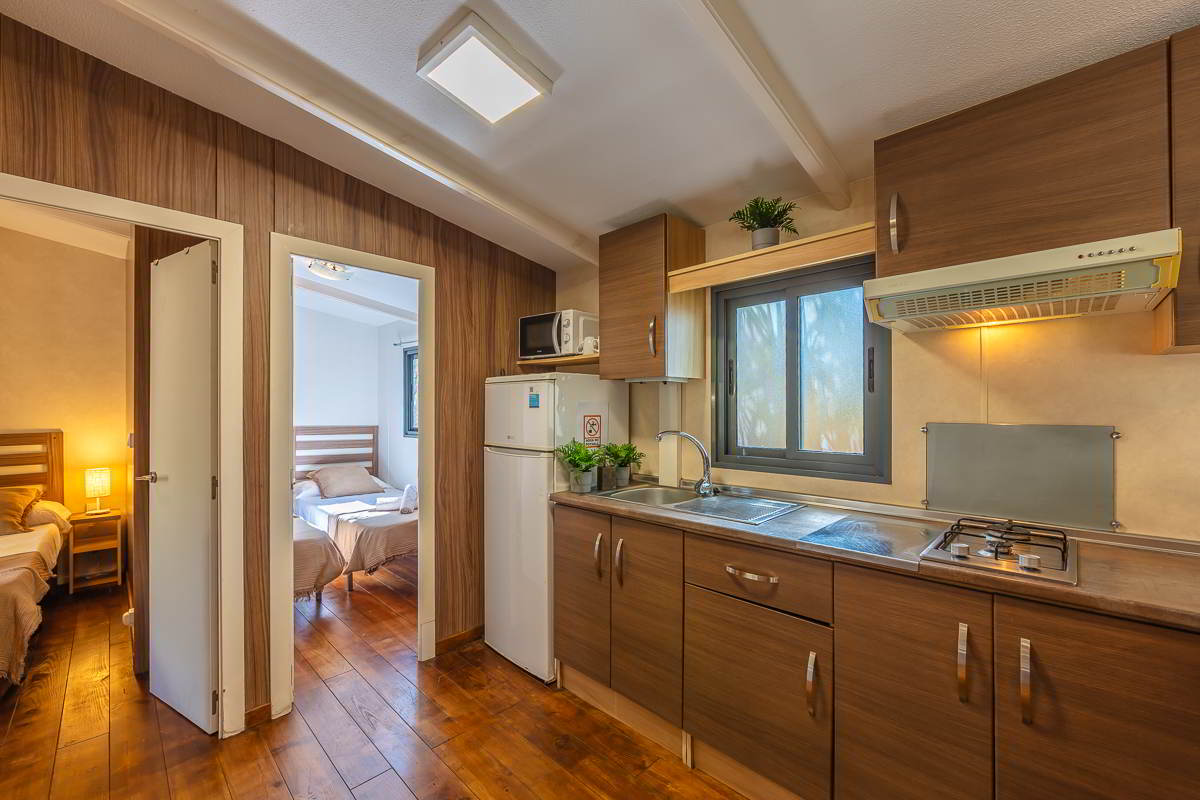 Cocina de la casa Nepal en Villas La Ahumada, con muebles en madera oscura, ventana con luz natural, encimera con fregadero y fogones, frigorífico, microondas y acceso directo a dos dormitorios con camas individuales y luz cálida