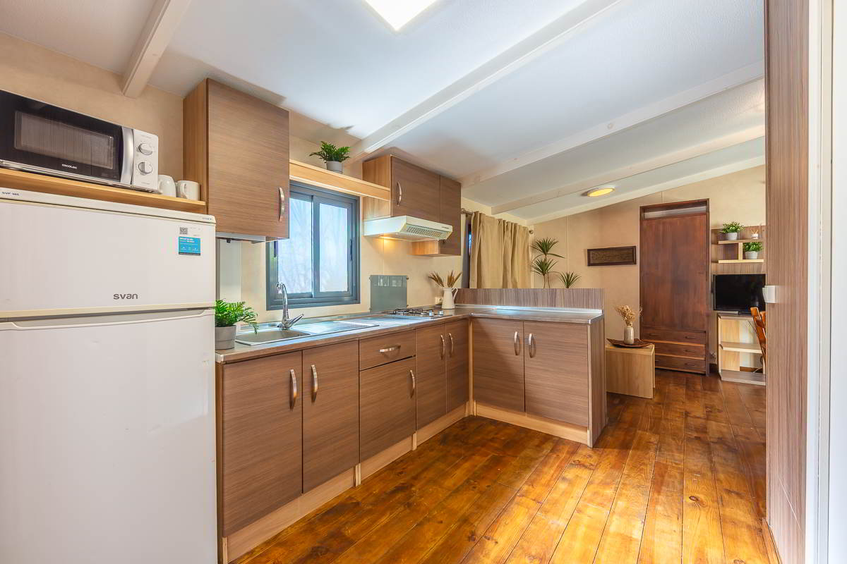 Vista general de la cocina y salón de la casa Nepal en Villas La Ahumada, con mobiliario de madera, encimera en forma de L, frigorífico blanco, microondas, ventana con luz natural, suelo de madera y zona de estar al fondo con televisión y decoración vegetal