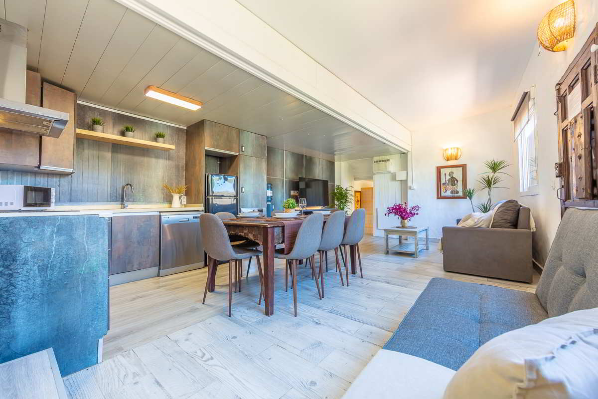 Cocina abierta con muebles oscuros de acabado natural, encimera clara, mesa de madera rústica con seis sillas modernas, salón con sofá, decoración con plantas y luz cálida en paredes blancas.