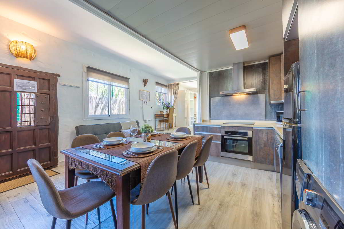 Espacio abierto con cocina moderna en tonos oscuros y comedor de seis plazas con mesa de madera y vajilla colocada, junto a puerta de entrada rústica y ventanas que aportan luz natural.