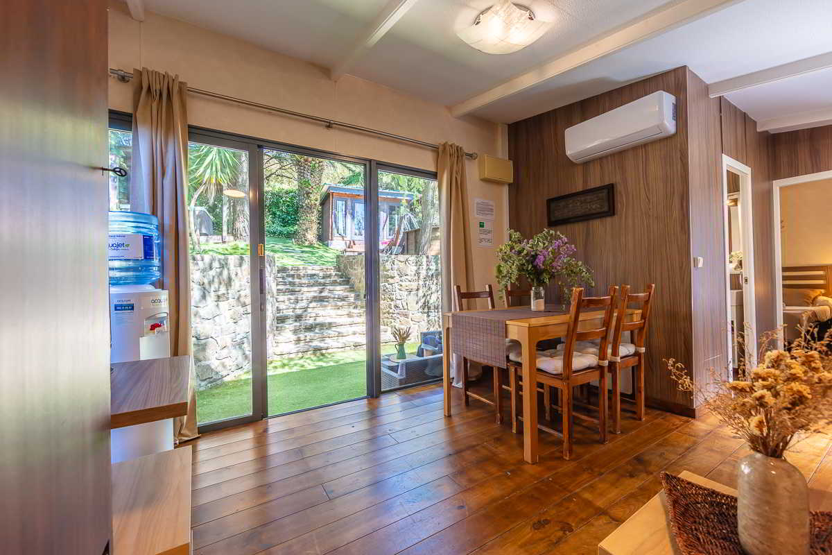 Comedor de la casa Nepal en Villas La Ahumada, con mesa de madera decorada con flores, suelo de madera natural, dispensador de agua a la izquierda y grandes ventanales con vistas al jardín escalonado y la casita exterior entre árboles