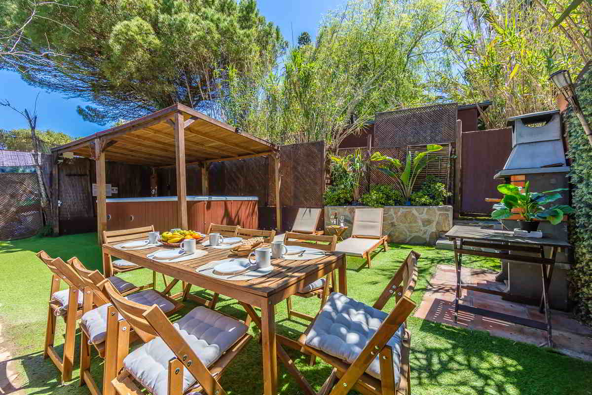 Jardín de Villa Verde con mesa de desayuno montada para seis personas, barbacoa, tumbonas y piscina exterior techada al fondo, rodeado de vegetación densa y árboles altos.