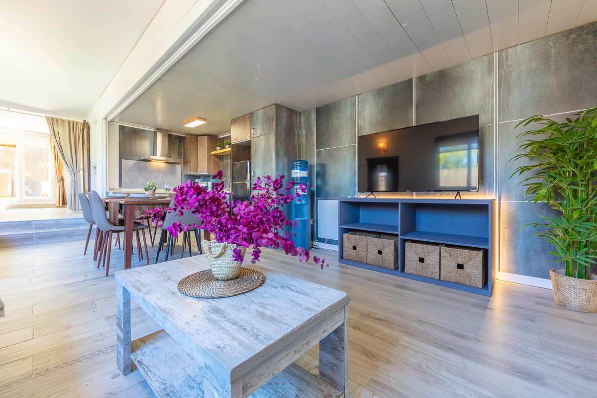 Vista desde el salón hacia la cocina y el comedor, con mesa de centro de madera blanca y jarrón con flores fucsias en primer plano, televisor sobre mueble azul con cestas, cocina con mobiliario en tonos oscuros y zona de comedor con sillas grises, todo iluminado por luz natural