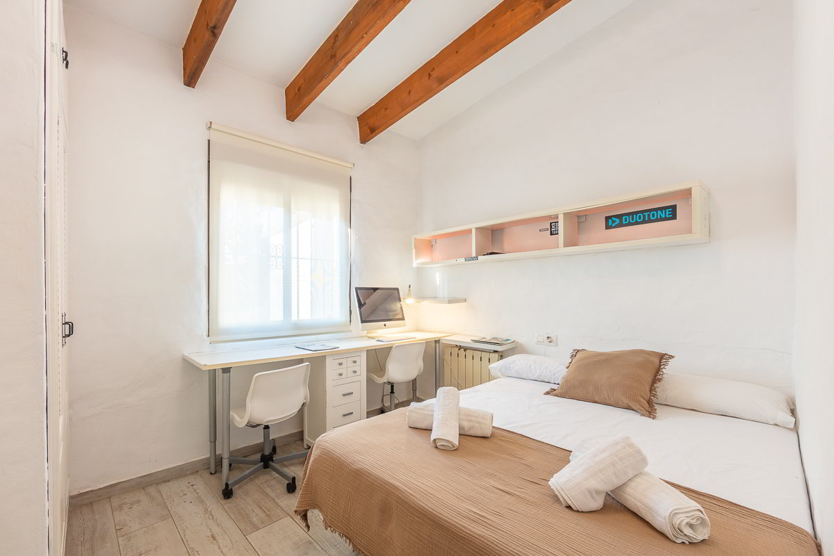 Dormitorio del Chalet La Ahumada con cama doble, escritorio en L con dos sillas, vigas de madera en el techo y luz natural suave.