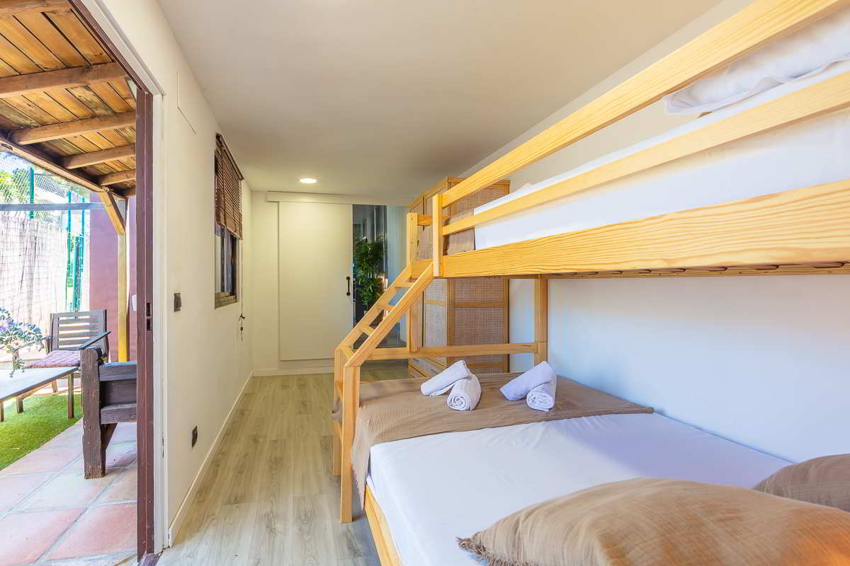 Litera de madera con cama doble inferior y cama individual superior en dormitorio luminoso con suelo de madera clara, cojines marrones, toallas enrolladas y acceso directo a un porche exterior con sillas de madera y suelo de terracota.