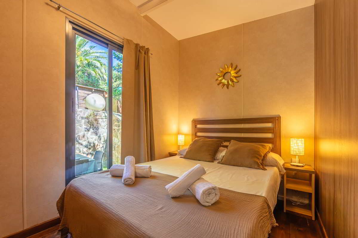 Dormitorio principal de la casa Nepal en Villas La Ahumada, con cama doble vestida en tonos tierra, cabecero de madera, lámparas de noche encendidas, decoración en forma de sol dorado en la pared y ventanal con vistas al jardín exterior