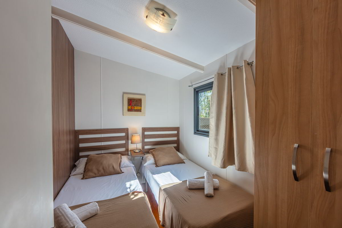 Dormitorio con dos camas individuales en la casa Nepal de Villas La Ahumada, con cabeceros de madera, lámpara de noche encendida, ventana con cortinas claras, armario de madera, decoración minimalista y luz natural