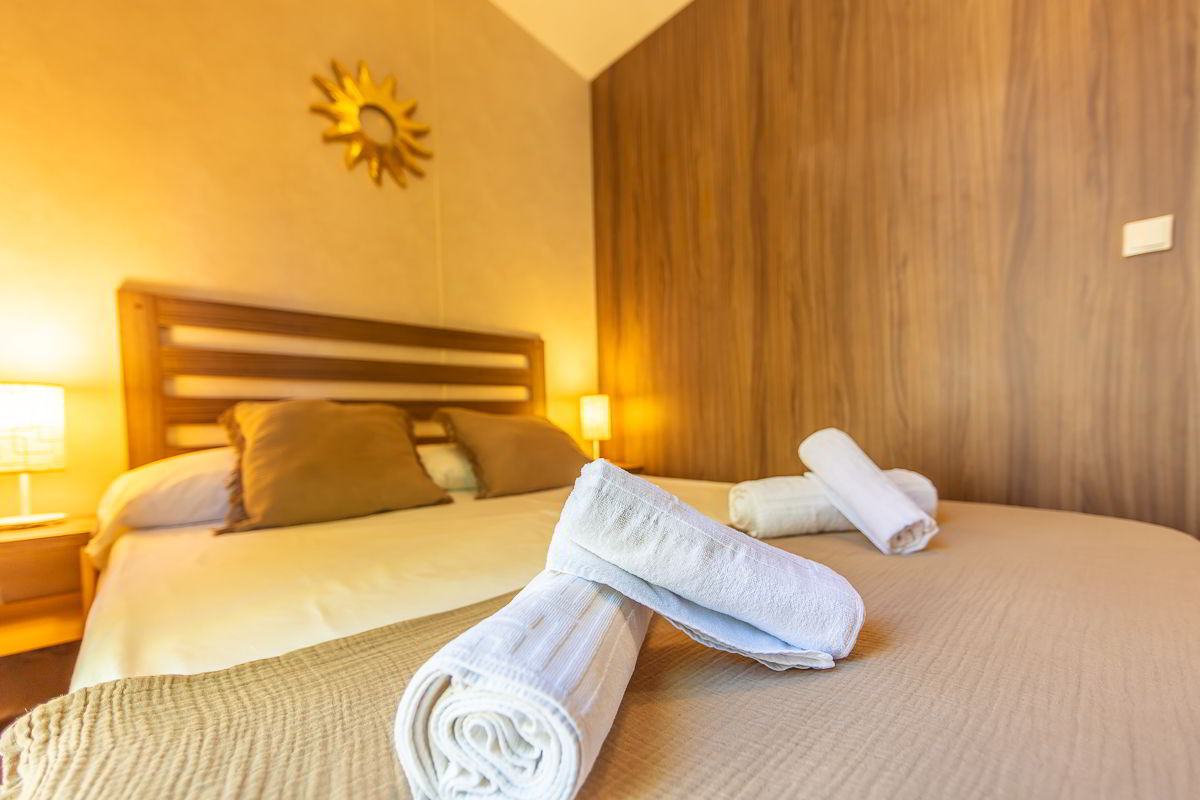 Detalle de la cama doble del dormitorio principal en la casa Nepal de Villas La Ahumada, con toallas enrolladas sobre la colcha beige, cojines marrones, cabecero de madera, lámparas encendidas y decoración dorada en forma de sol sobre la pared