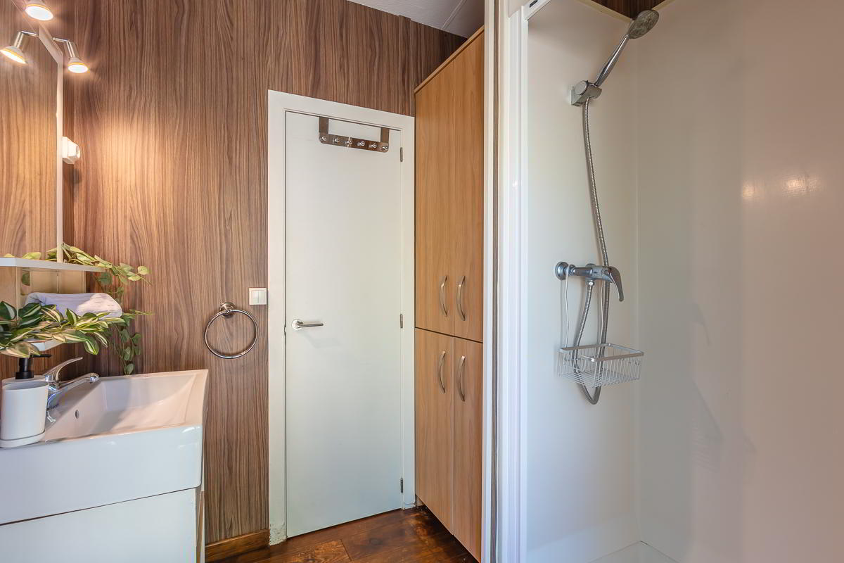 Baño de la casa Nepal en Villas La Ahumada, con ducha individual de pared blanca, grifería moderna, lavabo con estantería decorada con plantas y toallas, armario de almacenaje en madera clara y puerta blanca