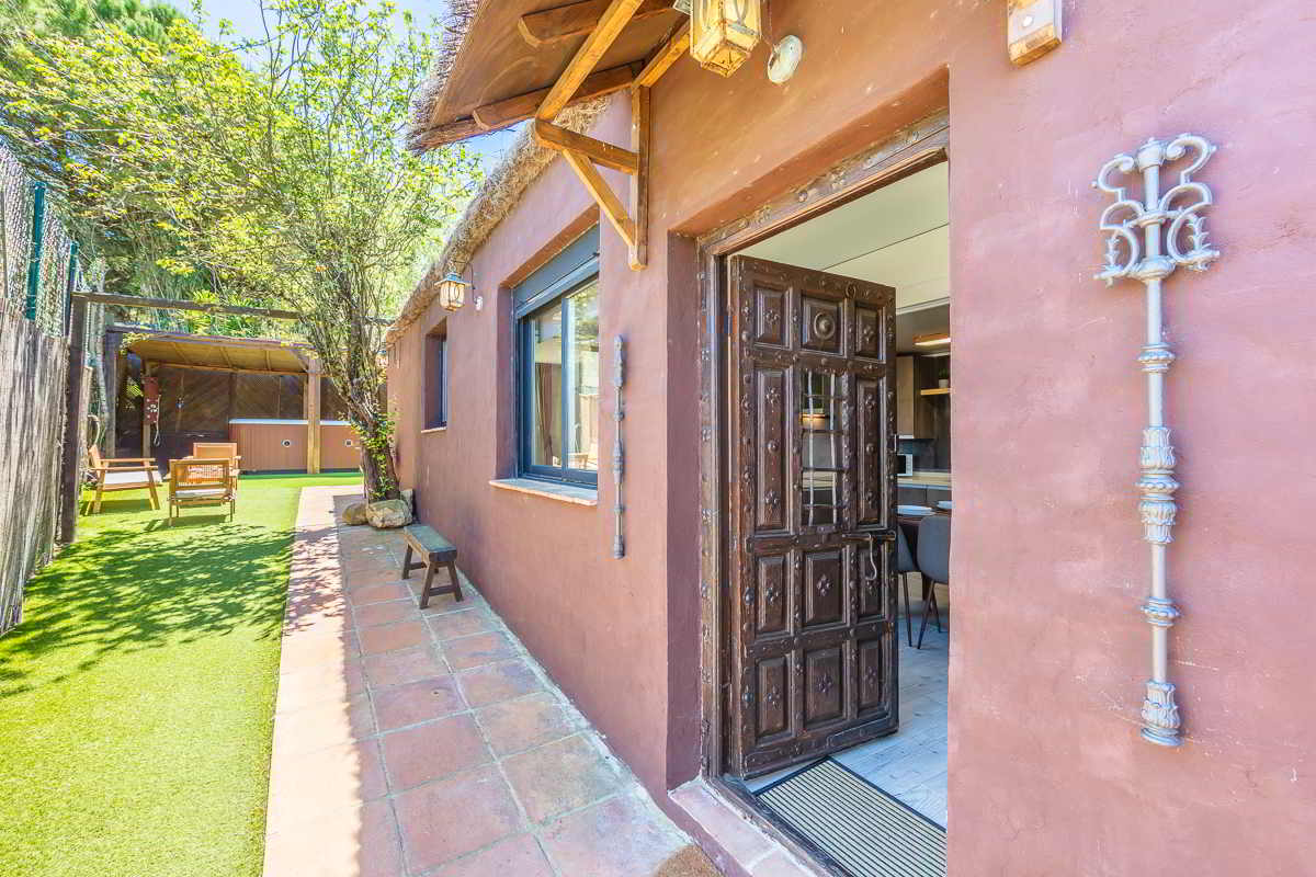 Fachada de una casa color terracota con puerta de madera maciza tallada, ventana amplia, faroles de hierro forjado, jardín con césped artificial, muebles de exterior bajo una pérgola de madera y sombra de árboles al fondo.