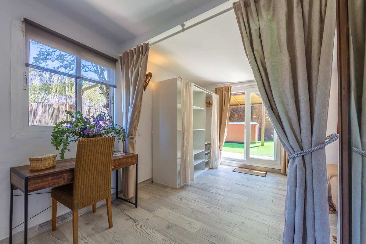 Espacio interior con escritorio de madera, silla de fibras naturales, estantería blanca con cortinas y grandes ventanales que se abren al jardín con jacuzzi exterior.