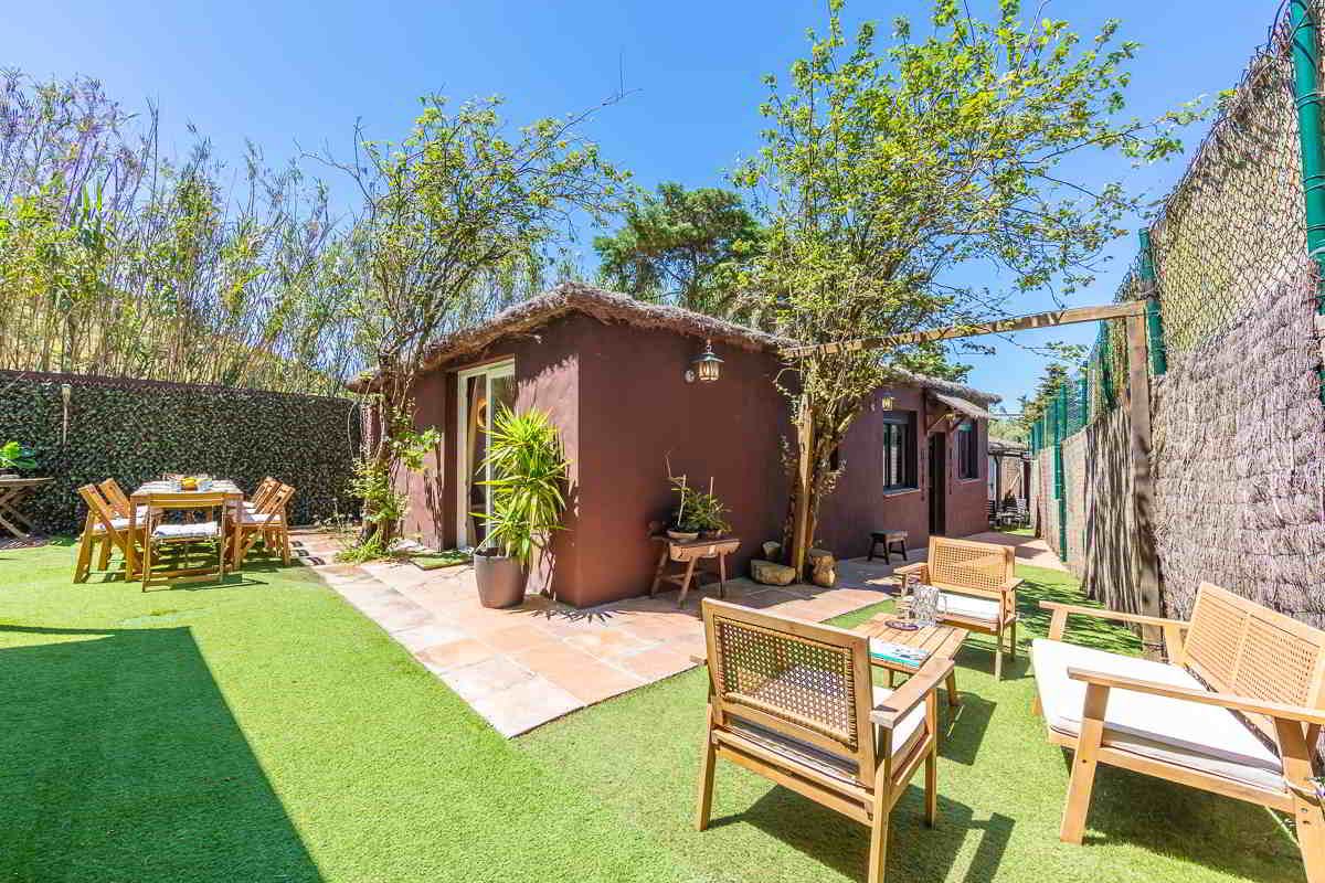 Vista del exterior de Villa Verde en Tarifa, con fachada color tierra, muebles de jardín en madera y una gran vegetación alrededor bajo un cielo azul.