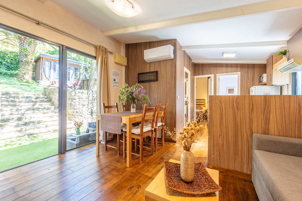 Vista interior de la casa Nepal en Villas La Ahumada, con comedor de madera decorado con flores frescas, suelo de madera, cocina integrada al fondo, puertas abiertas a los dormitorios y gran ventanal con vistas al jardín y casita exterior