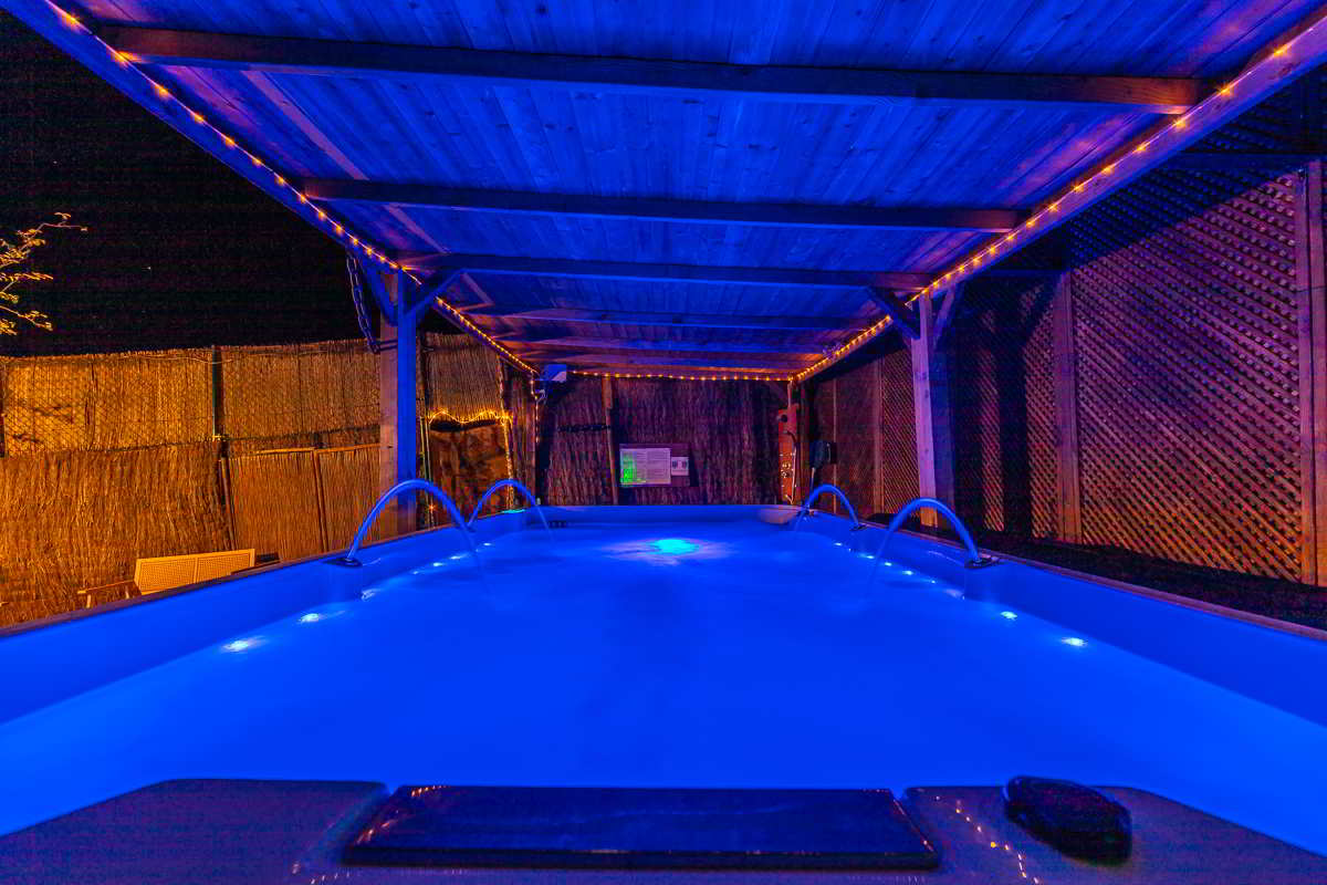 Vista frontal de un jacuzzi iluminado en azul, cubierto por una pérgola de madera con luces cálidas, rodeado por paneles de cañizo y paredes de madera, creando un ambiente íntimo y relajante.