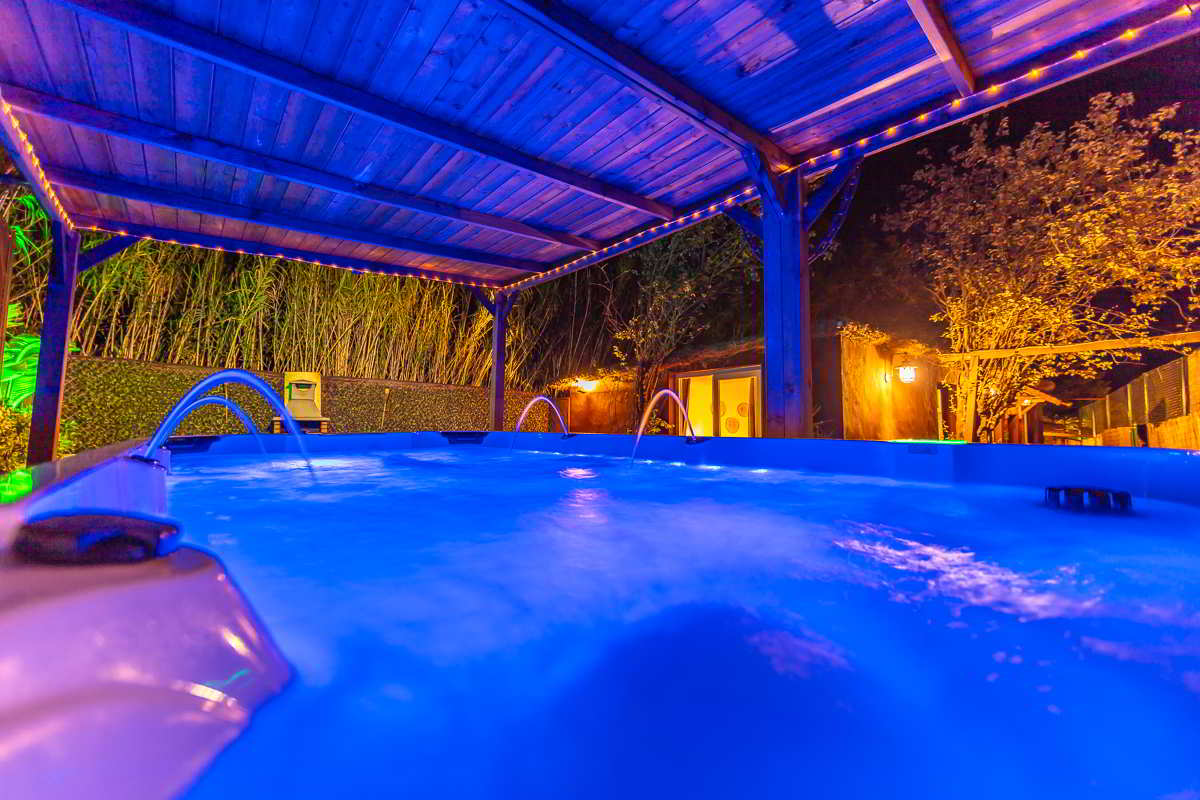 Vista desde el interior del jacuzzi iluminado en azul con surtidores encendidos, bajo un porche de madera con guirnaldas cálidas, rodeado de cañaverales y árboles iluminados, con la entrada a la casa al fondo decorada con luz tenue y ambiente acogedor.