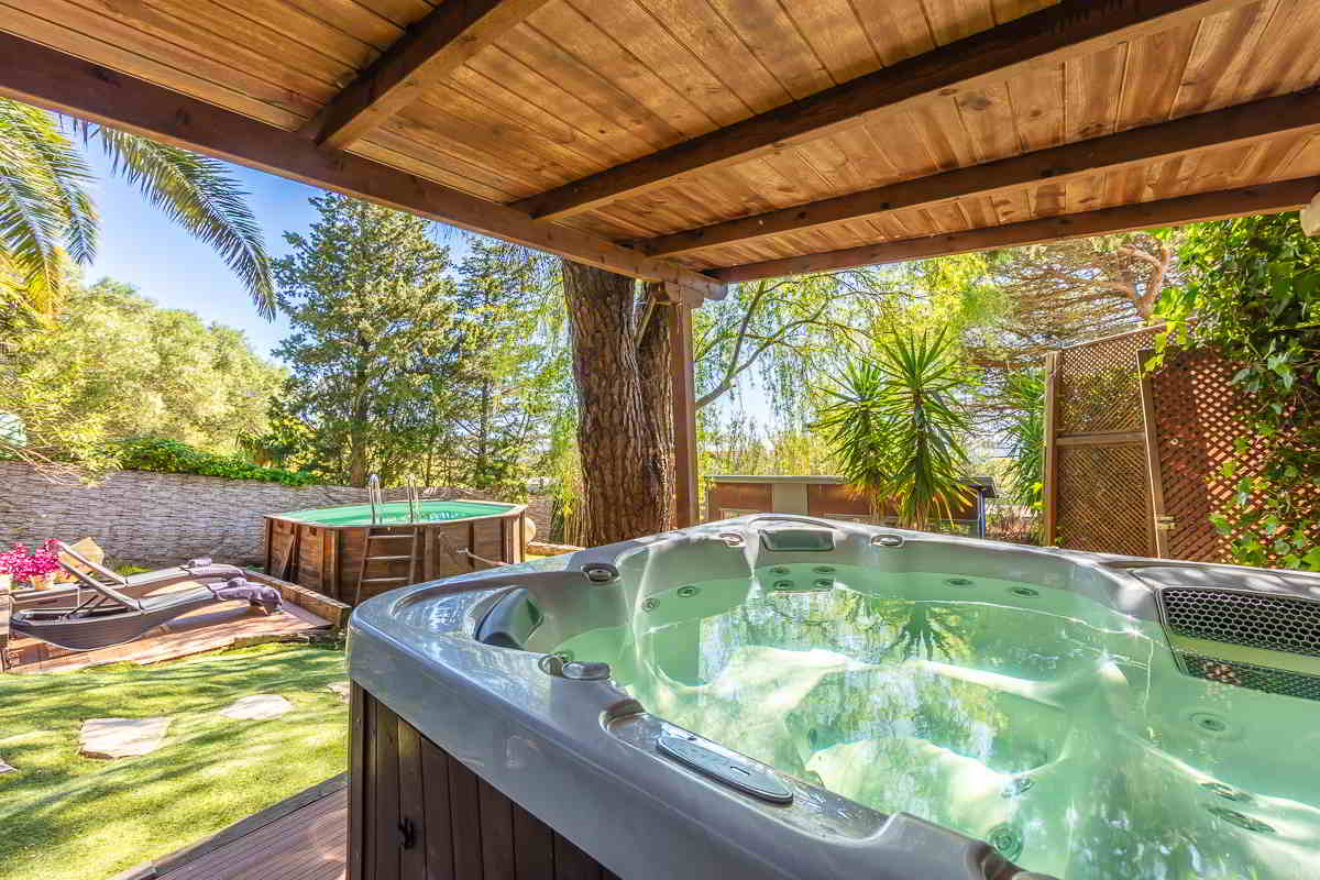 Jacuzzi cubierto con estructura de madera, rodeado de vegetación y vistas a una piscina redonda elevada, tumbonas de madera y jardín con césped artificial bajo la sombra de grandes árboles.