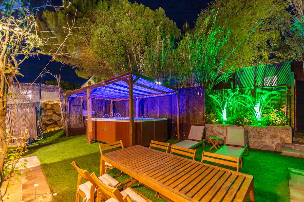 Jardín iluminado de noche con jacuzzi bajo pérgola de madera, rodeado de luces tenues, zona de tumbonas, mesa de comedor al aire libre y vegetación con focos verdes junto a una pared de piedra.