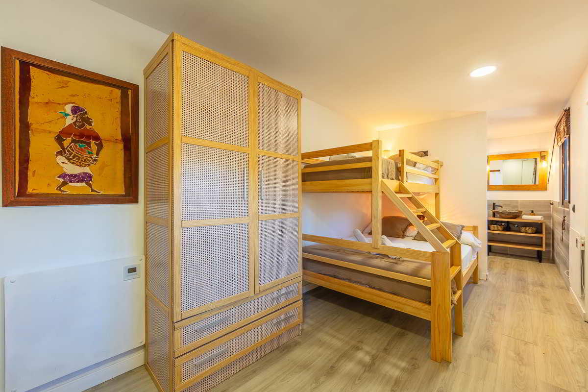 Dormitorio con litera de madera clara, cama inferior de matrimonio y cama superior individual, armario trenzado a juego, suelo de madera, iluminación cálida y acceso directo a baño con lavabo doble y espejo de madera.