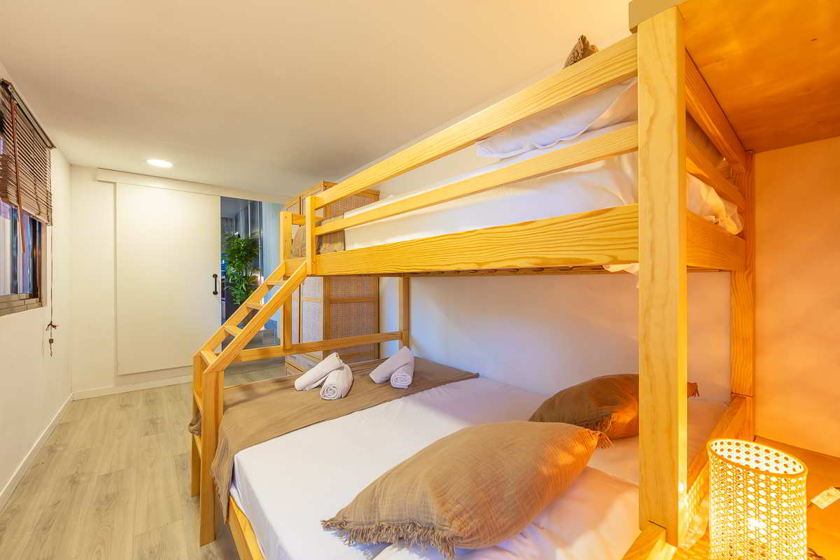 Detalle de una litera de madera con cama doble inferior y cama individual superior, ropa de cama en blanco y marrón, cojines decorativos, lámpara de mimbre encendida y puerta corredera blanca al fondo junto a planta decorativa