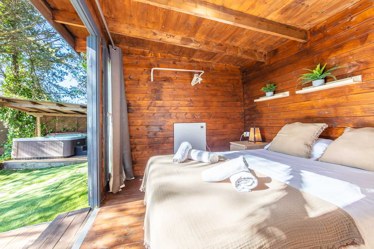 Habitación de madera cálida con cama doble vestida en tonos tierra, toallas enrolladas sobre la colcha, estanterías con plantas decorativas, lámparas de mesita y puerta abierta con acceso al jardín, donde se ve un jacuzzi cubierto entre árboles y césped.