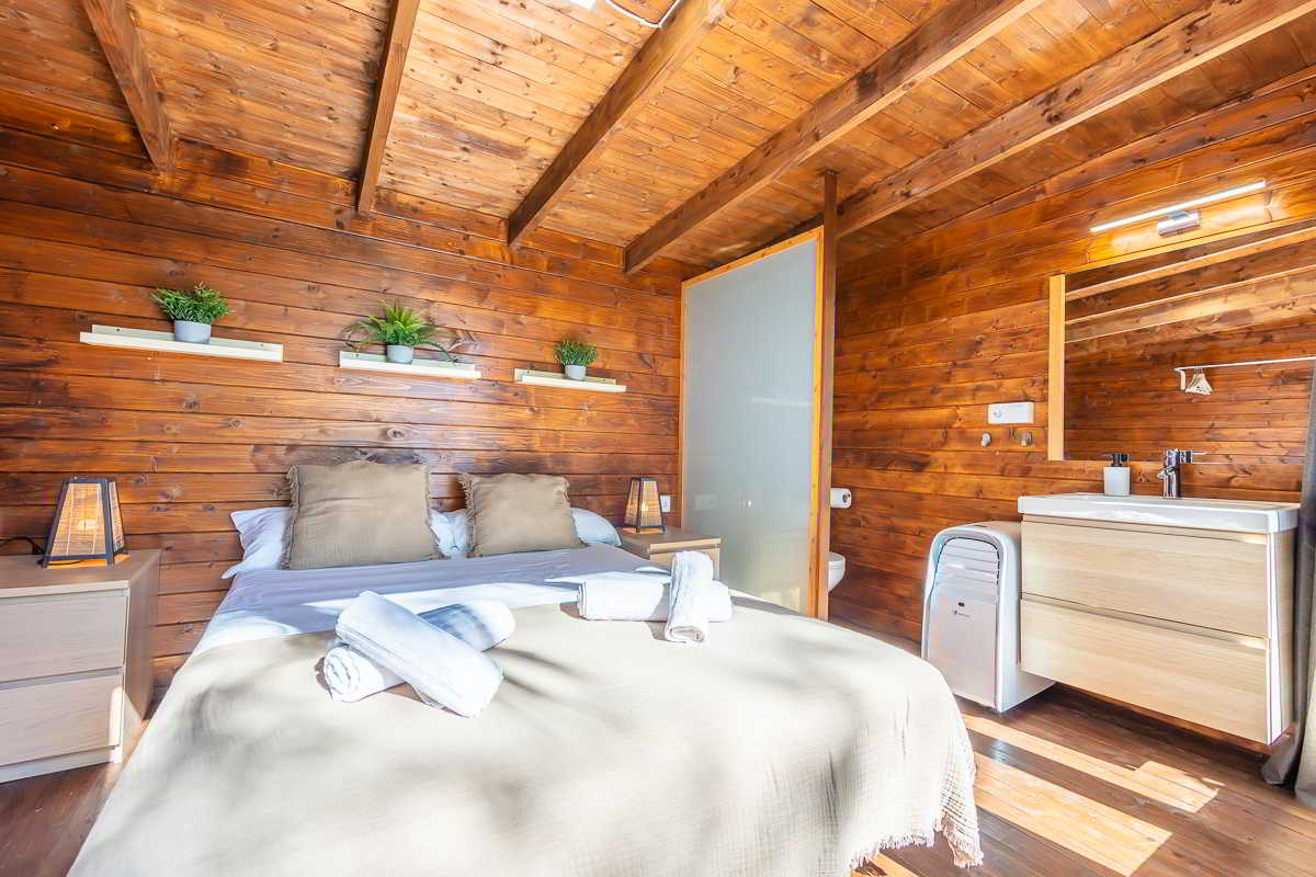 Interior de una habitación de madera con techo a dos aguas y vigas vistas, cama doble con manta beige, cojines marrones y toallas enrolladas, flanqueada por dos mesitas con lámparas cálidas, estanterías con plantas sobre el cabecero, y baño integrado con lavabo, espejo amplio y mampara de cristal esmerilado.