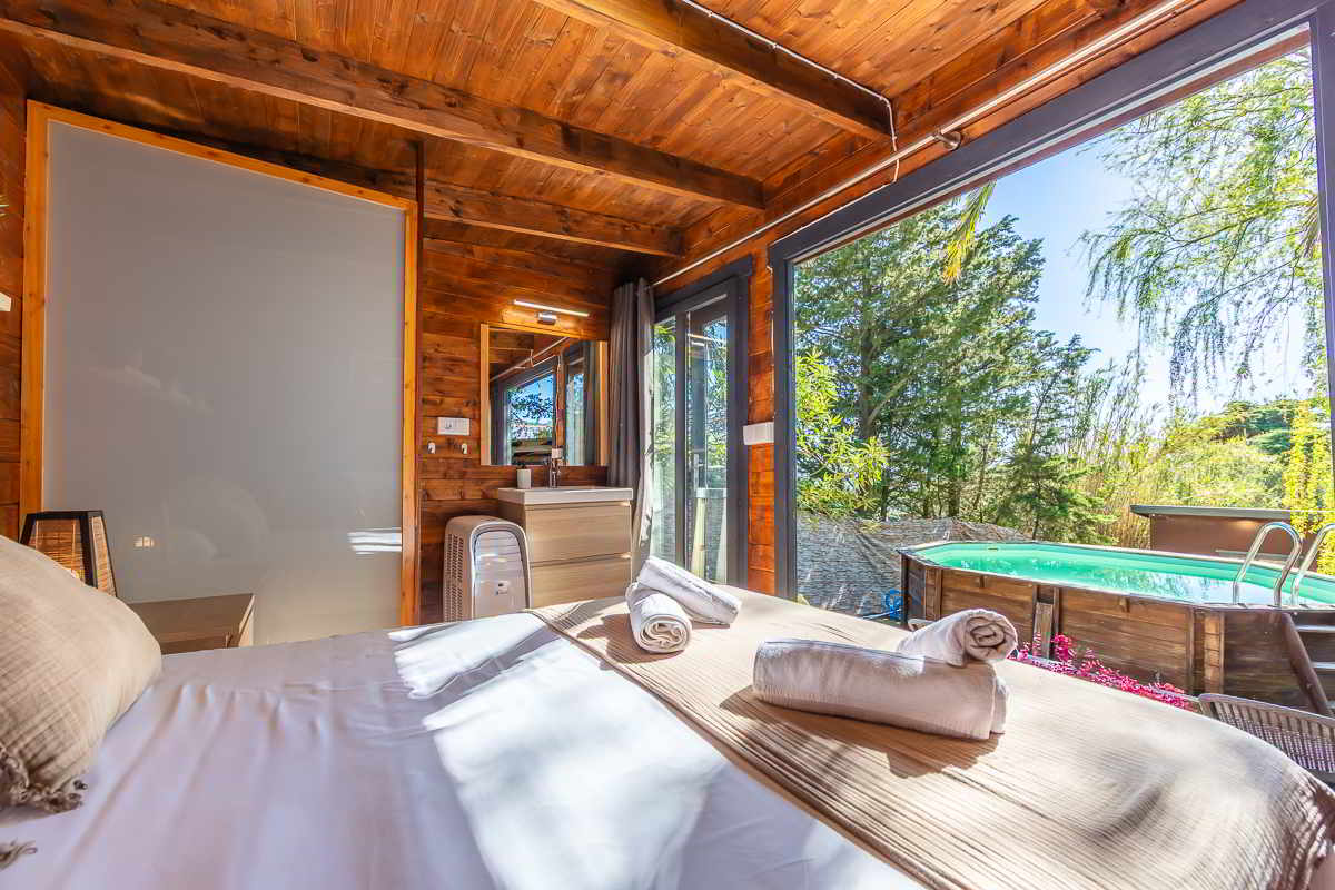 Vista desde la cama de una habitación de madera con techos de vigas, toallas enrolladas sobre una manta beige, lavabo con espejo y mueble claro, mampara translúcida a la izquierda, grandes ventanales abiertos que dan acceso directo a una terraza con piscina elevada rodeada de naturaleza.