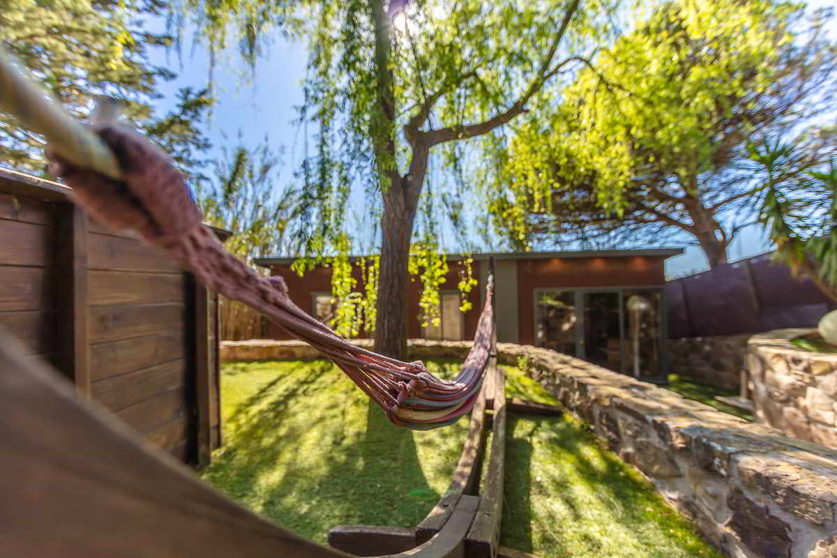 Primer plano desenfocado de una hamaca de cuerdas sujetada entre estructuras de madera, con vista al fondo del jardín rodeado de árboles, un sauce llorón y un edificio de fachada marrón con cristaleras.