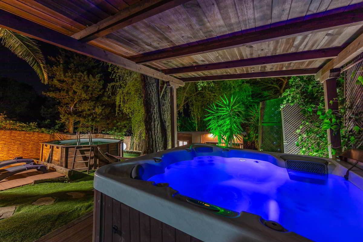 Jacuzzi iluminado con luz azul bajo un porche de madera, en un jardín rodeado de árboles y vegetación, con piscina elevada de madera y tumbonas en el fondo, todo ambientado con luces nocturnas cálidas y verdes.