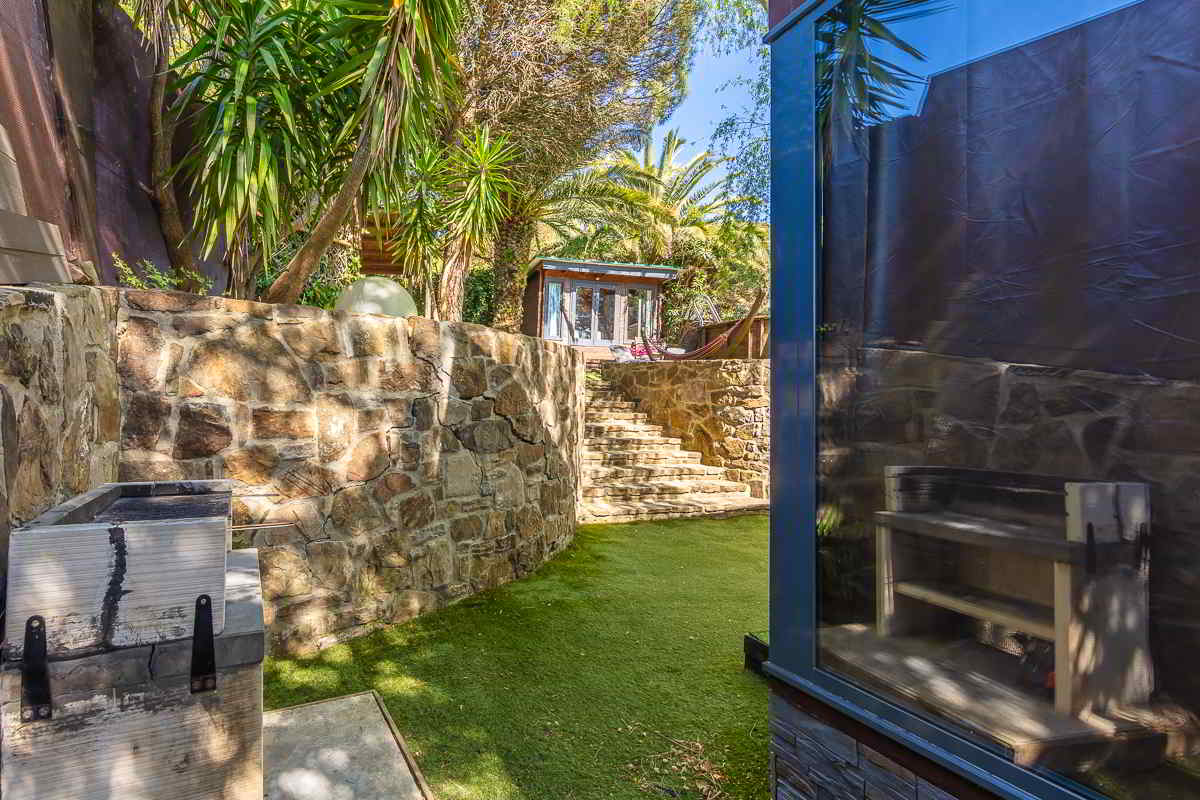 Vista parcial del jardín de la casa Nepal en Villas La Ahumada, con escalera de piedra, vegetación exuberante, zona de barbacoa en primer plano y estructura acristalada que refleja los árboles