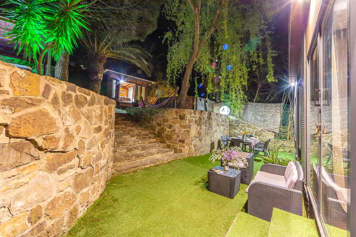 Vista nocturna de un jardín con césped artificial rodeado por muros de piedra, con escaleras que suben hacia una cabaña iluminada, zona de hamacas junto a la piscina, y un área de comedor exterior con mesa y sillones de mimbre bajo un árbol colgante y luces cálidas.