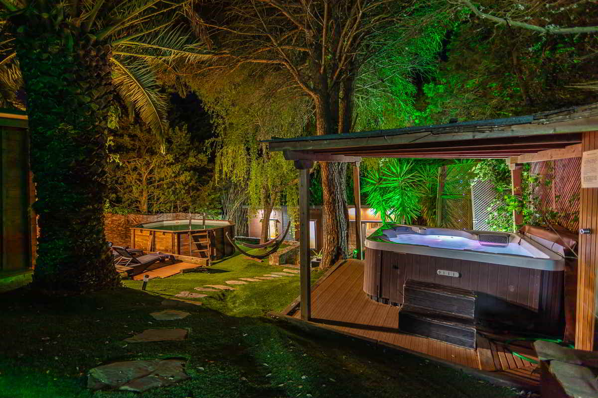 Vista nocturna de un jardín rodeado de árboles iluminados, con camino de piedras que lleva a una zona de relax con jacuzzi cubierto de madera, piscina elevada también de madera, tumbonas sobre tarima, hamaca colgante y exuberante vegetación tropical.