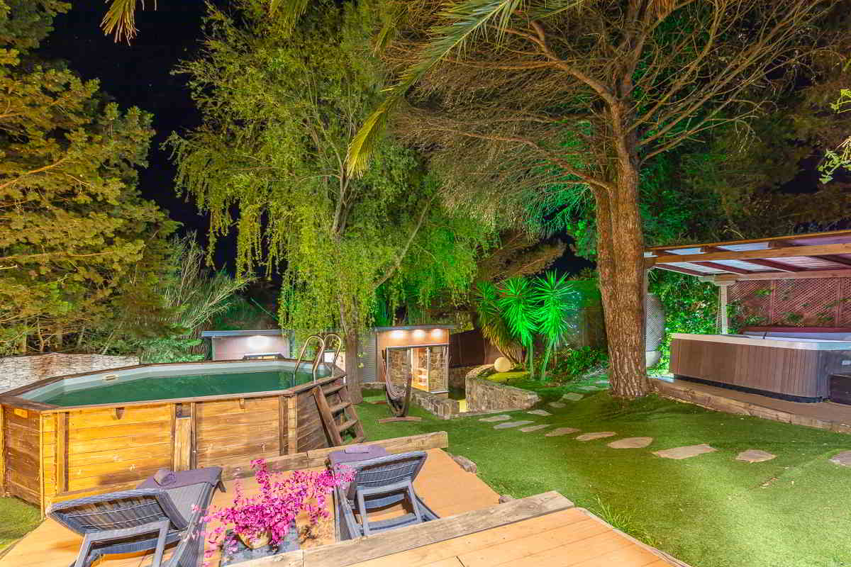 Vista nocturna de un jardín rodeado de árboles y plantas iluminadas, con piscina elevada de madera, tumbonas con toallas sobre tarima, hamaca colgante entre árboles y un jacuzzi cubierto al fondo bajo pérgola de madera.