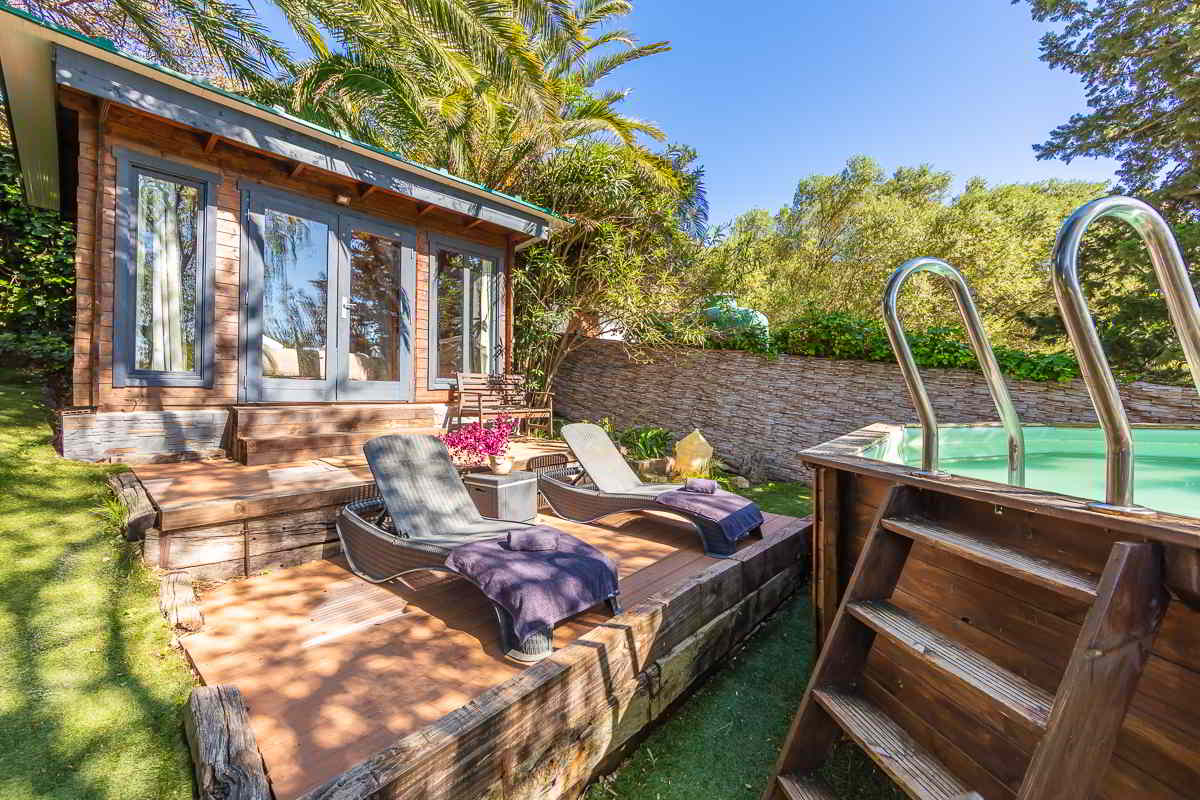 Vista del jardín del alojamiento con piscina elevada de madera, escalerilla metálica, tumbonas con toallas, flores fucsias y una casita de madera rodeada de vegetación exuberante y palmeras altas, todo bajo un cielo despejado.