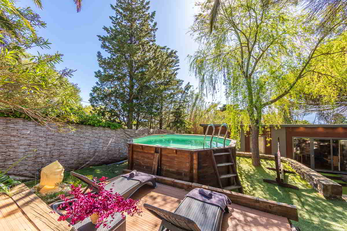 Piscina elevada de madera rodeada de césped artificial, tumbonas de mimbre con toallas, flores decorativas y un busto de Buda en un jardín íntimo y rodeado de naturaleza en Tarifa.