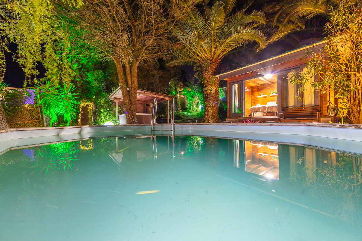 Vista nocturna desde el interior de una piscina elevada, rodeada de árboles, palmeras y luces ambientales, con una cabaña iluminada al fondo y un jacuzzi cubierto parcialmente visible entre la vegetación.