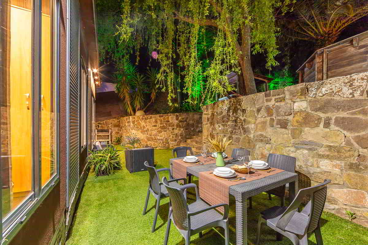 Terraza nocturna con césped artificial junto a una fachada moderna, con una mesa de comedor exterior preparada para seis personas, iluminación cálida, decoración vegetal, y un muro de piedra que rodea el espacio, bajo un gran árbol con ramas colgantes y luces ambientales.