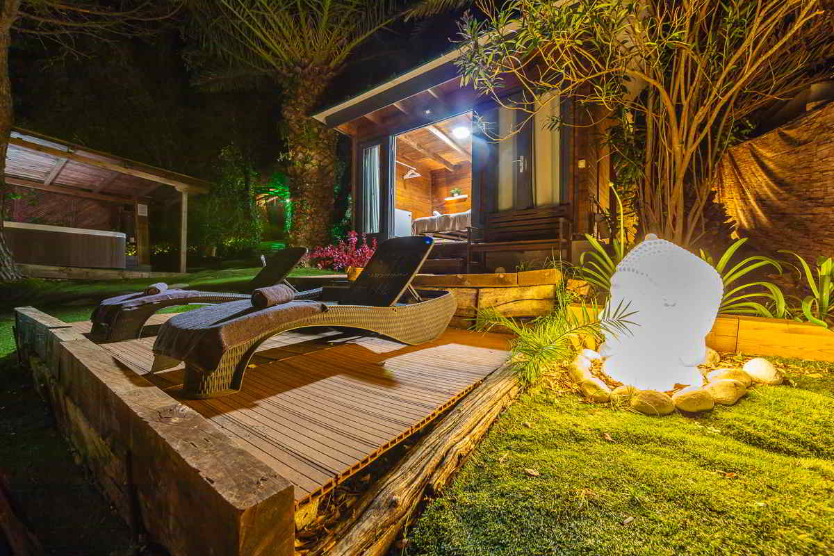 Vista nocturna de una zona de relax con dos tumbonas modernas sobre una tarima de madera, toallas enrolladas, una figura de Buda iluminada sobre piedras y césped, y una cabaña de madera cálidamente iluminada al fondo.