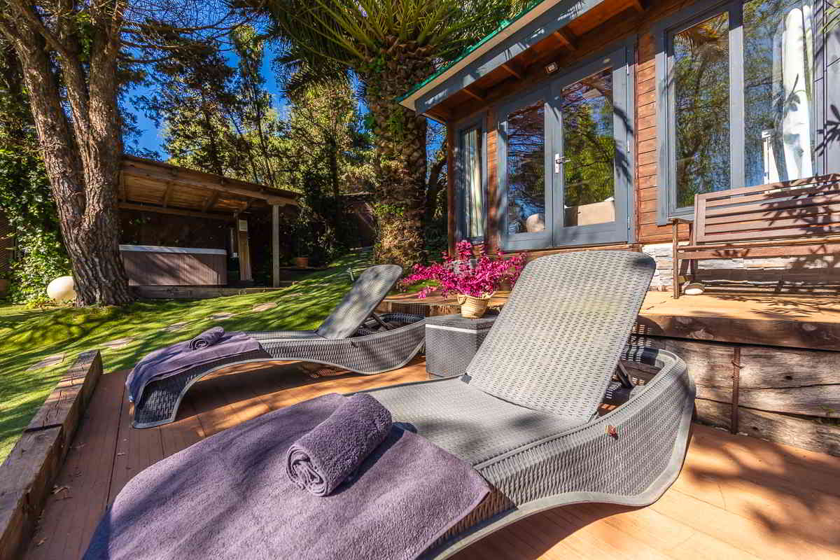 Primer plano de dos tumbonas de ratán con toallas moradas en una tarima de madera, situadas frente a una cabaña de madera con puertas de cristal, junto a una jardinera con flores fucsias y un banco de madera; al fondo, un jacuzzi cubierto entre árboles.