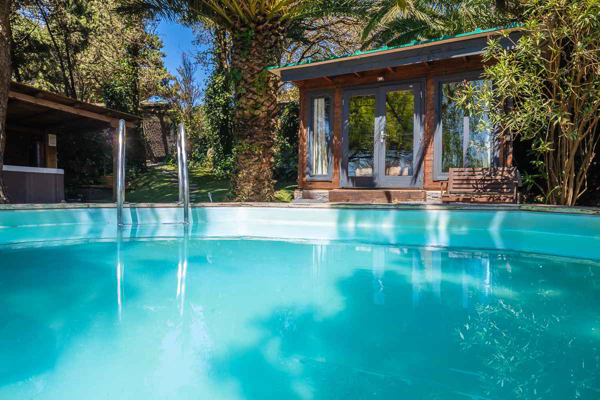 Vista de la piscina de la casa Nepal en Villas La Ahumada, rodeada de palmeras y vegetación frondosa, con escalerilla metálica, casita de madera con grandes ventanales al fondo y banco de madera junto a la entrada