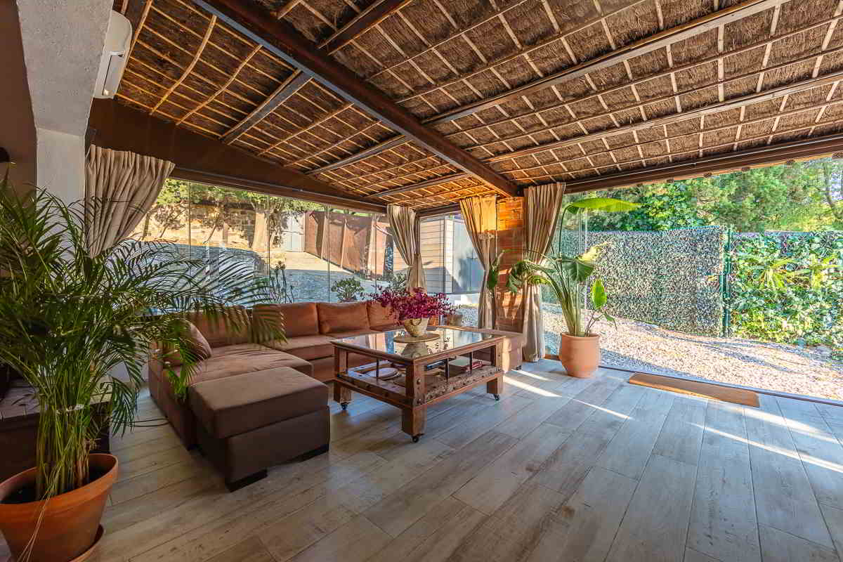 Porche cerrado del chalet La Ahumada con techo de cañizo, cortinas recogidas, sofá chaise longue de terciopelo marrón y mesa de centro de madera, rodeado de vegetación y luz natural.