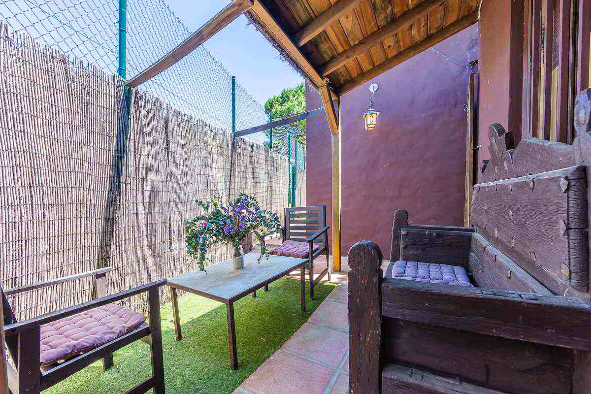 Detalle del porche exterior con techo de madera, lámpara rústica de pared, mesa baja decorada con un jarrón de flores moradas, y bancos de madera oscura con cojines, sobre suelo de césped artificial y baldosas.